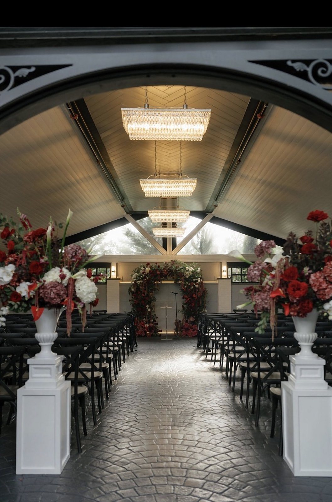 Aisle view through an archway to a floral ceremony arch beneath tiered crystal chandeliers